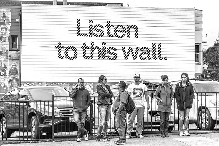 People standing in front of a billboard with the text "Listen to this wall" 